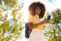 From below of glad ethnic female in earphones and sunglasses taking selfie on cellphone under blue sky in sunlight — Photo de stock