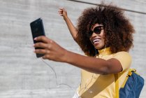 Feliz mujer étnica en auriculares y gafas de sol tomando selfie en el teléfono celular cerca de la pared gris a la luz del sol - foto de stock