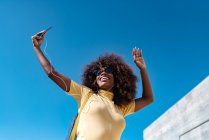 Von unten eine fröhliche ethnische Frau mit Kopfhörern und Sonnenbrille, die unter blauem Himmel im Sonnenlicht ein Handy-Selfie macht — Stockfoto
