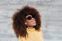 Молодая веселая этническая женщина с африканской прической слушает музыку и смеется на солнце возле серой стены — стоковое фото