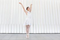 Young female ballet dancer in tiptoe in pointe shoes with raised leg and arm dancing on tiled pavement outdoors — Stock Photo
