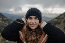 Улыбающаяся молодая женщина надевает тёплую шляпу и смотрит в камеру, стоя на грубом высокогорье в пасмурный день. — стоковое фото