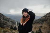 Smiling young female putting on warm hat and looking at camera while standing on rough highlands on cloudy gloomy day — Stock Photo