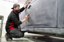 Seitenansicht des männlichen Meisters in schmutziger Arbeitskleidung, der das Auto säubert, während er das Fahrzeug für die Lackierung im Reparaturservice vorbereitet — Stockfoto
