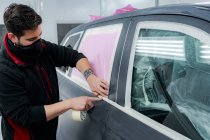 Umtriebiger Mechaniker in Maske klebt Klebeband auf Auto vor Lackierung in Werkstatt — Stockfoto