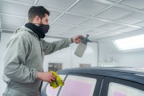 Vista lateral do trabalhador masculino em máscara e pintura uniforme automóvel ao usar pistola de pintura — Fotografia de Stock