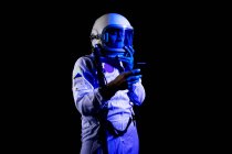 Созерцательный мужчина-космонавт в белом скафандре и шлеме, просматривающий современный смартфон, стоя на черном фоне — стоковое фото