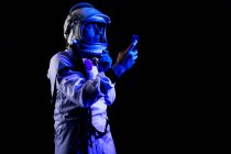 Созерцательный мужчина-космонавт в белом скафандре и шлеме, просматривающий современный смартфон, стоя на черном фоне — стоковое фото