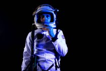 Hombre cosmonauta con traje espacial blanco y casco mientras está de pie sobre fondo negro en luz de neón azul mirando a la cámara - foto de stock