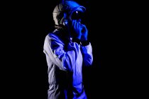Side view of male cosmonaut wearing white space suit and helmet while standing on black background in blue neon light looking away — Stock Photo