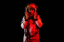 Male cosmonaut wearing white space suit and helmet while standing on black background in red neon light looking at camera — Fotografia de Stock