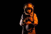 Contemplative male cosmonaut in white spacesuit and helmet browsing modern smartphone while standing on black background — Fotografia de Stock