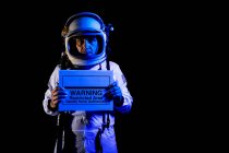 Cosmonauta macho adulto serio en traje espacial y casco demostrando pancarta con texto Advertencia Área restringida Fuerza mortal Autorizado mientras está de pie sobre fondo negro y mirando a la cámara - foto de stock