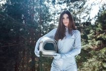 Жіночий космонавт в космосі тримає шолом в руках і стоїть в засніженому лісі, дивлячись на камеру — стокове фото