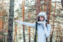 Astronauta joven enfocada en traje espacial y casco mirando hacia otro lado de pie en un bosque nevado con el brazo extendido - foto de stock