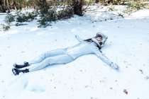 Pieno corpo in forma calma donna spaziale in costume e casco sdraiato con le braccia tese su radura innevata nella foresta invernale — Foto stock