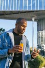 Adult ethnic African American male with bottle of orange juice surfing internet on cellphone in city — Stock Photo
