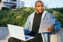 Empleado remoto masculino étnico adulto con documento y netbook sentado en la ciudad mientras mira hacia otro lado - foto de stock