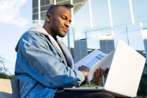 From below adult ethnic male remote employee with document and netbook sitting in city — Stock Photo