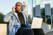 Ethnic African american adult male remote employee working on laptop while speaking on mobile phone sitting in city — Stock Photo