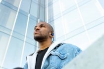 From below of adult bearded ethnic male in wireless earphones listening to song while looking away in town — Fotografia de Stock