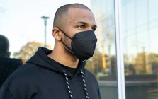 Varón barbudo adulto con máscara respiratoria y sudadera con capucha negra mirando hacia adelante contra la pared de vidrio durante la pandemia de COVID 19 en la ciudad - foto de stock