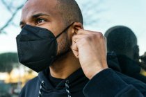 Adult bearded ethnic male in respiratory mask and black hoodie looking forward against glass wall during COVID 19 pandemic in town — Fotografia de Stock