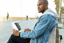 Vista lateral de un empleado remoto masculino adulto afroamericano étnico con computadora portátil sentado en la ciudad - foto de stock