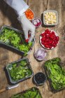 Зверху врожаю анонімний професійний шеф-кухар в рукавичці додавання стиглих червоних вишневих помідорів до свіжих змішаних листя у фользі, розміщених на столі біля салату овочевих інгредієнтів — стокове фото