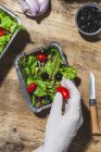 From above crop anonymous professional chef in glove adding ripe red cherry tomatoes to fresh mixed leaves in foil bowl placed on table near salad vegetable ingredients — Stock Photo