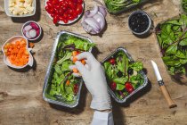 From above crop anonymous professional chef in glove adding carrot slices in foil bowl placed on table near salad vegetable ingredients — Fotografia de Stock