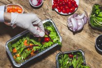 From above crop anonymous professional chef in glove adding onion slices to fresh mixed leaves in foil bowl placed on table near salad vegetable ingredients — Fotografia de Stock