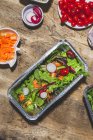 Top view composition of delicious vegetable salads in foil bowls placed on table near various ingredients including cherry tomatoes onions radish and carrots — Fotografia de Stock