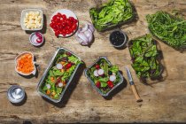 Top view composition of delicious vegetable salads in foil bowls placed on table near various ingredients including cherry tomatoes onions radish and carrots — Fotografia de Stock