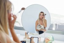 Очаровательная женщина сидит с котом перед зеркалом и наносит порошок, делая макияж дома — стоковое фото