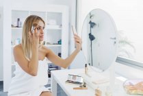 Vista lateral de la mujer con la piel perfecta aplicando polvo y tocando la cara mientras toma uno mismo tiro en el teléfono inteligente mientras hace maquillaje en casa - foto de stock