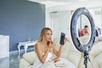 Vista laterale della femmina che applica lucidalabbra e si guarda allo specchio mentre è seduta in camera con lampada a cerchio — Foto stock