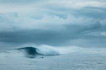 Unrecognizable person swimming on surfboard on powerful foamy sea waves rolling and splashing over water surface against cloudy blue sky — Fotografia de Stock