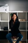 Cheerful young Hispanic woman wearing black jacket and jeans sitting on chair in modern light room and looking away — Fotografia de Stock