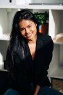 Cheerful young Hispanic woman wearing black jacket and jeans sitting on chair in modern light room and looking at camera — Fotografia de Stock
