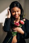 Joven modelo femenino étnico encantador con bata negra tocando el cabello mientras mira el ramo de rosas rosadas en la sombra de la luz del sol - foto de stock