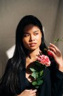 Young charming ethnic female model wearing black robe touching hair while looking away holding pink roses bouquet in shadow from sunlight — Photo de stock