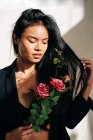 Young charming ethnic female model wearing black robe touching hair while looking at pink roses bouquet in shadow from sunlight — Fotografia de Stock