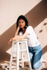 Full body of confident young Hispanic lady wearing casual clothes standing on knees and leaning on wooden chair in shadow from sunlight — Photo de stock