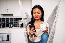 Молода іспаномовна жінка сидить у гамаку на сучасній кухні з гарячим напоєм і користується сучасним смартфоном вдень. — стокове фото