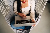 From above of crop smiling young woman with long dark hair using touchpad of portable laptop with earphones while sitting in hammock indoors — Fotografia de Stock