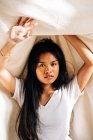 Attractive young Hispanic woman wearing white t shirt touching blanket with raised hands while looking at camera in sunshine — Photo de stock