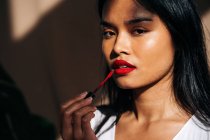 Portrait of crop thoughtful ethnic female with long dark hair looking at camera and rouging lips with red lipstick — Stock Photo