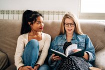 Young wistful female friends in casual clothes sitting on comfortable sofa and reading book while spending time together — Stock Photo