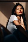 Attractive young Hispanic woman wearing white cloth touching face while looking away in sun shadow — Photo de stock